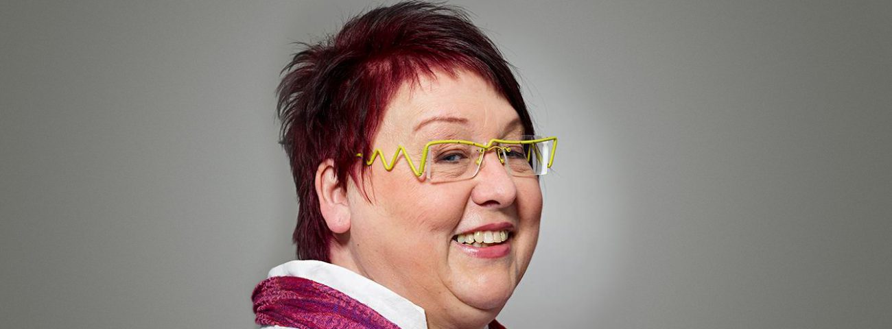 Fröhliche Frau mit kurzen roten Haaren und gelber Brille schaut von links nach rechts.